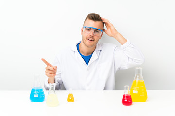 Young scientific with lots of laboratory flask surprised and pointing finger to the side