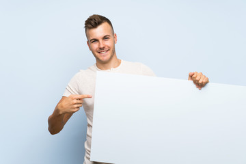 Young handsome blonde man over isolated blue background holding an empty white placard for insert a...