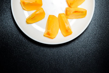 Persimmon fruit ripe small pieces in white plate top over black background.