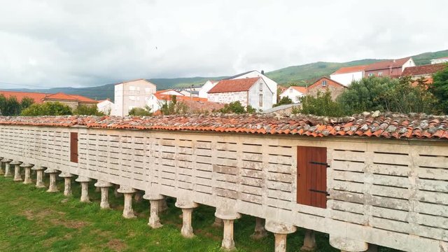 Drone footage of Horreo de Carnota in Vilar, A Coruna, Spain on a bright cloudy day with green grass in the foreground and spanish style houses in the background