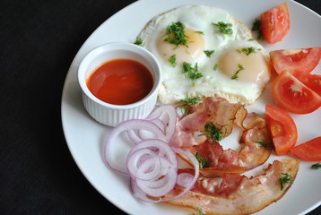 Fried eggs with tomatoes, red onion, bacon for breakfast on a white plate on the black background. Delicious homemade breakfast.