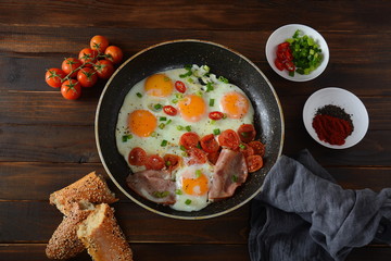 Fried eggs in a pan with cherry tomatoes, bacon and bread for breakfast on a wooden background. Delicious breakfast concept.
