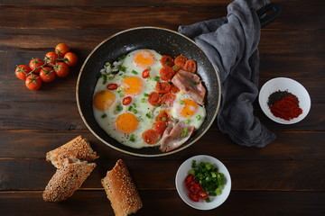 Fried eggs in a pan with cherry tomatoes, bacon and bread for breakfast on a wooden background. Delicious breakfast concept.