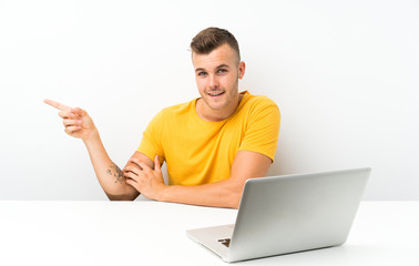 Young blonde man in a table with a laptop pointing finger to the side