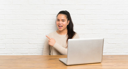 Young teenager Asian girl with a laptop in a table pointing finger to the side