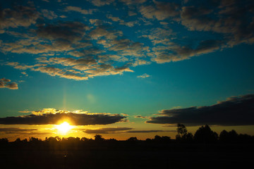 Obraz premium Sunset, darkness and night, view to the sun through dark clouds on bright blue sky, black silhouettes of trees. High contrast. Australia.