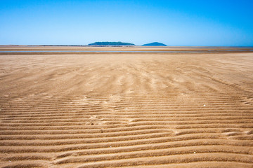 The coastal strip (coastline, beach) at low tide. A wide strip of desert sand to the horizon, a desert area, a wavy relief runs along it. An island is in the centre. Makey, Queensland, Australia.