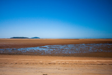 The coastal strip at low tide. A wide strip of desert sand to the horizon, a desert area with wet sand. An island's mountains on the horizon at the left. Makey, Queensland, Australia.