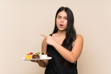 Young teenager Asian girl holding lots of different mini cakes over isolated background surprised and pointing side