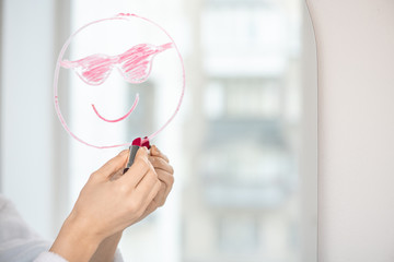 Hand of girl with lipstick drawing face with smile and sunglasses on mirror