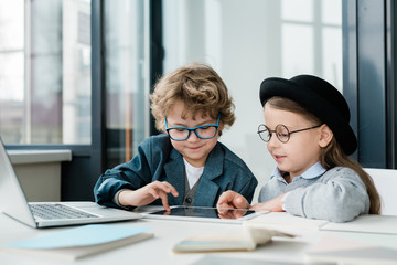 Cute elementary school friends in casualwear and eyeglasses using digital tablet