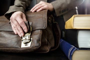 Hand fastens a buckle on an old brown, classic, leather briefcase, which stands on a table, next to a stack of books.