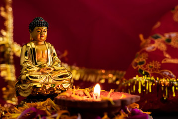 beautiful small statue of buddha in maditation position with shining diya on flower petals against red golden background. religion and tarditional concept.jpg