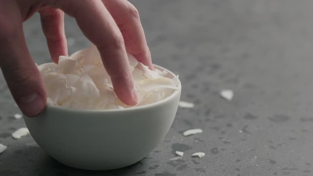 Slow motion man hand takes coconut chips from white bowl on terrazzo surface