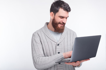 Smiling young bearded man is looking through a computer and smiling near white background.