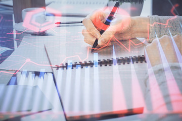 Hand taking notes in notepad. Forex chart holograms in front. Concept of research. Double exposure