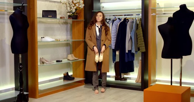 a brunette with long hair is in the fitting room. the woman is dressed in a brown knee-length coat and classic trousers. she picks up and tries on beige high-heeled shoes