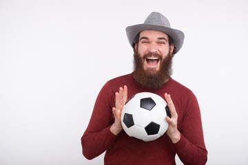A young happy bearded man is holding with both hands a soccer ball near a white wall.