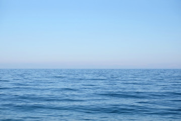 Calm sea surface with small waves in the morning and the land on skyline in light haze in pink and blue colors under clear cloudless sky horizontal view