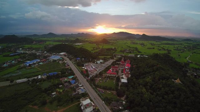 VDO. Aerial view evening above green rice paddy field, houses and buddhist temple with mountains and cloudy sky background, sunset at Wat Khao Chong Pran (Bat Temple), Photharam, Ratchaburi, Thailand.