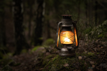 Vintage gasoline oil lantern lamp burning with a soft glow light in an dark forest / wood. Light in...