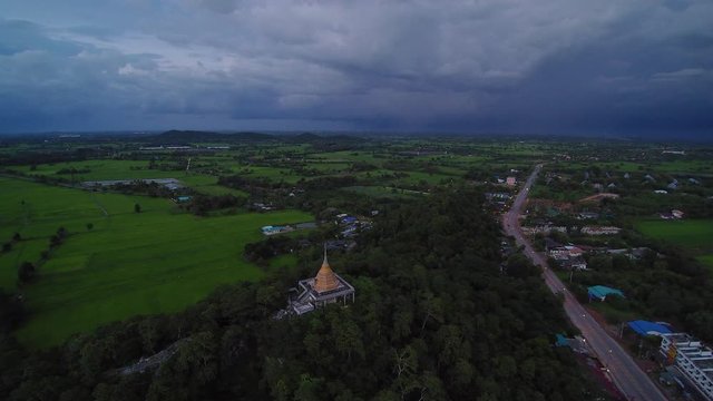 VDO. Aerial view panning of golden pagoda on top hill and long road with green paddy fields and rainy sky background, sunset with rain at Wat Khao Chong Pran (Bat Temple), Ratchaburi, Thailand.