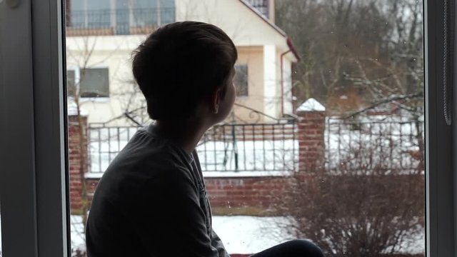 child kid sitting on the window sill and looking the window on falling snowflakes. snowing winter landscape. bored, sick boy wants to go outdoor to play with snow.home quarantine isolation virus