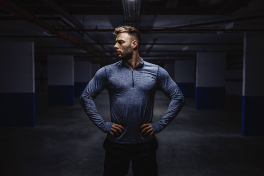 Powerful Strong Caucasian Man In Sportswear Standing In Garage With Hands On Hips And Looking Away. Nighttime. Urban Life Concept.