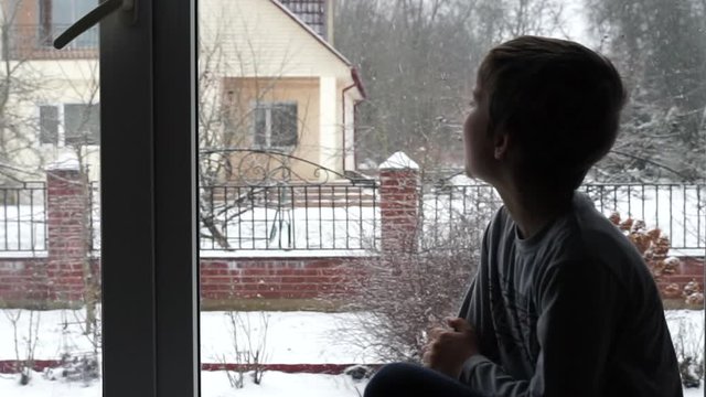 happy child enjoing snow weather. kid sitting on the window sill and looking through the window on falling snowflakes. snowing winter landscape.