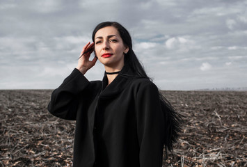  portrait of a woman in a black coat whose hair is flying in the wind stands in a field