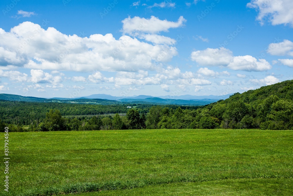 Poster Green field surrounded by short trees and mountains on a sunny day