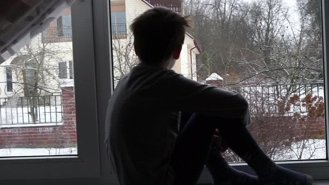  quarantine time at School. home isolation from virus. child kid sitting on the window sill and looking through the window on falling snowflakes. sick boy wants to go outdoor to play