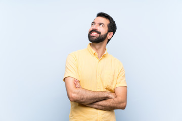 Young man with beard over isolated blue background looking up while smiling