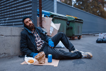 Dirty beggar with food sitting at the trashcan