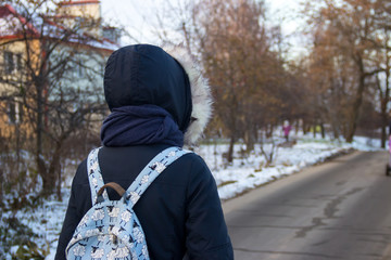 The girl in a blue jacket with a hood, scarf and blue backpack walking along the road, looking into the distance. End of autumn, early winter. On the side of the road is snow and trees