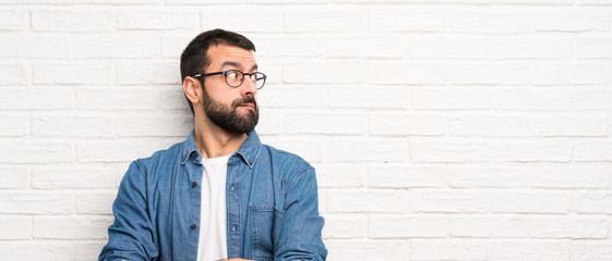 Handsome man with beard over white brick wall with confuse face expression