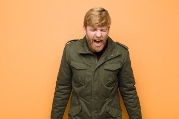 young blonde man shouting aggressively, looking very angry, frustrated, outraged or annoyed, screaming no against flat wall