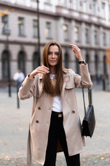 Fototapeta premium Outdoor photo of brunette lady walking on street background in autumn day.Fashion street style portrait. wearing dark casual trousers, white sweater and creamy coat.Fashion concept.