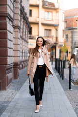 Outdoor photo of brunette lady walking on street background in autumn day.Fashion street style portrait. wearing dark casual trousers, white sweater and creamy coat.Fashion concept.