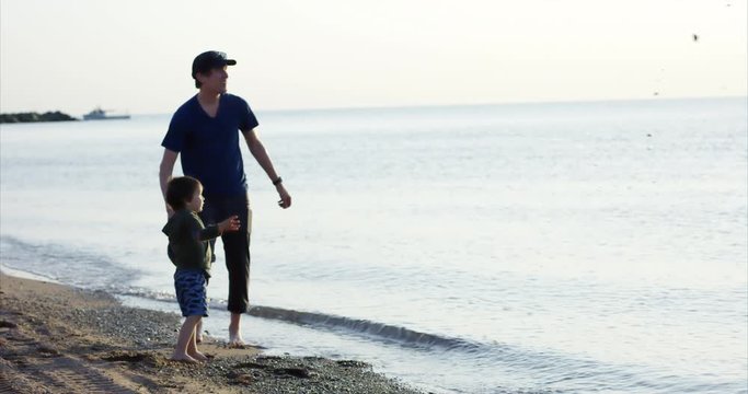 Father and toddler son throwing rocks into ocean at sunset - wide shot