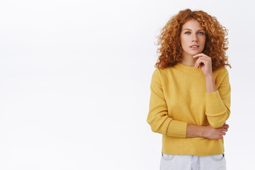 Thoughtful, serious-looking redhead curly woman in yellow sweater have interesting thought, touch chin and squinting as see something interesting, pondering what to do, stand white background