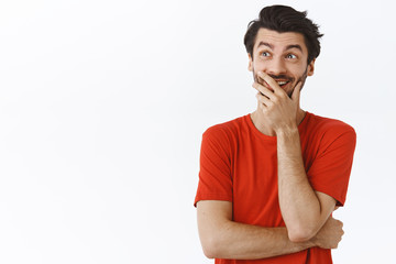 Enthusiastic young man with beard, messy hairstyle, wear red t-shirt, laughing out loud, chuckling and cover mouth as hiding smile, look upper left corner, imaging something hilarious