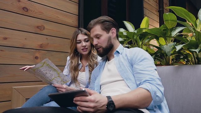 Good-looking elated modern young woman and man which sitting on soft couch in hotel hall and using tablet pc and map