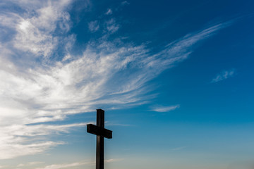 Silhouette of a simple oak wood catholic cross against dramatic blue sky with white clouds.