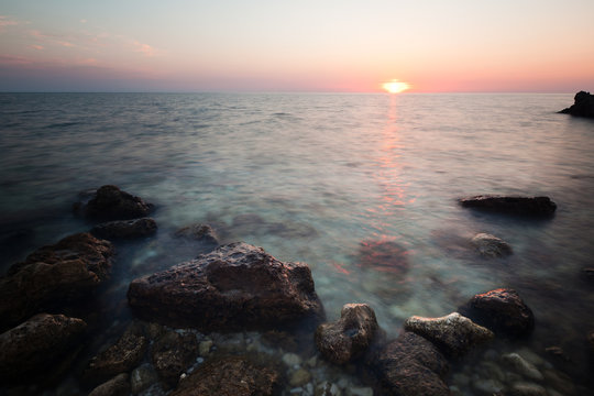 Seascape of still sea waters shore, rocky coastline and romantic pink sunset
