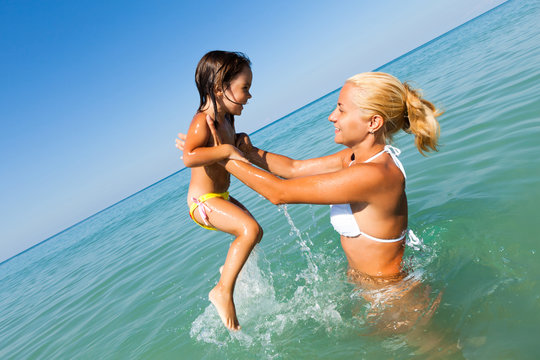Young blond mother in white bikini standing in still blue sea water and playing with her small smiling daughter on sunny summer day. Family vacations and travelling concept