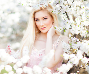 Young beautiful blonde smiling woman in white mini dress standing with blooming trees at background on clear sunny day. Woman natural beauty and summer nature concept