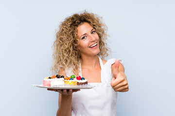 Young blonde woman with curly hair holding lots of different mini cakes with thumbs up because something good has happened