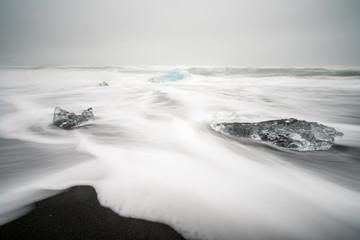 Fototapeta na wymiar Crystal clear and blue ice chunks washes up on the black lava sand by the waves on diamond beach in Jokulsarlon glacier lagoon. Climate change and glacier melting concept.