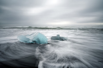 Crystal clear and blue ice chunks washes up on the black lava sand by the waves on diamond beach in Jokulsarlon glacier lagoon. Long exposure shot.